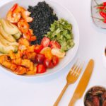 a bowl of black rice, avocado, tomatoes, shrimps, sweet potatoes with chilli peppers on the side on a white table with cutlery