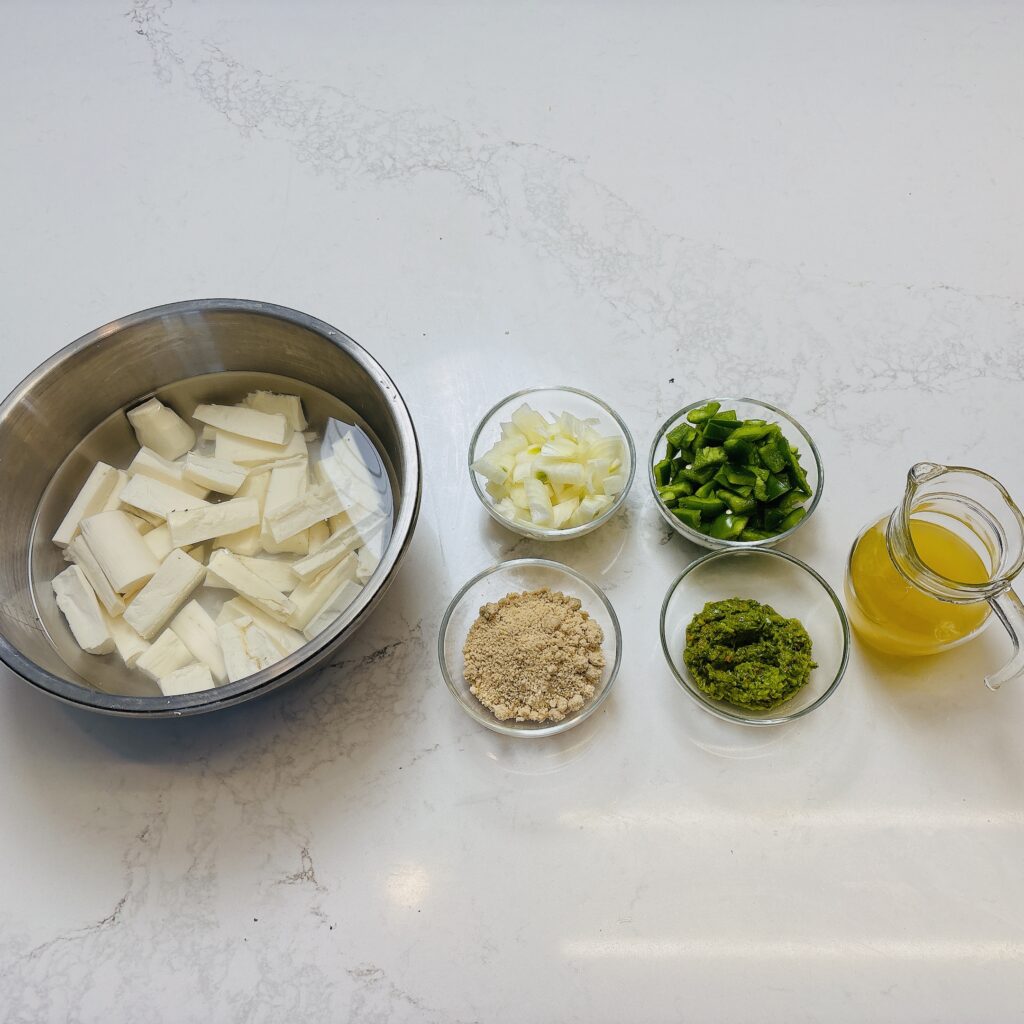 cassava peeled and cut in a bowl, chopped green peppers in bowl, egusi powder in bowl, egusi seasoning in bowl, chicken broth in bowl.
