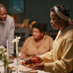 Black woman serving roasted turkey on dining table with floral decorations while family enjoying festive holiday dinner at home