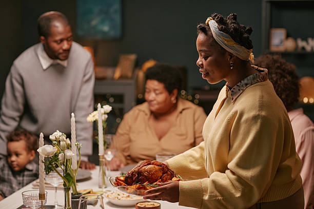 Black woman serving roasted turkey on dining table with floral decorations while family enjoying festive holiday dinner at home