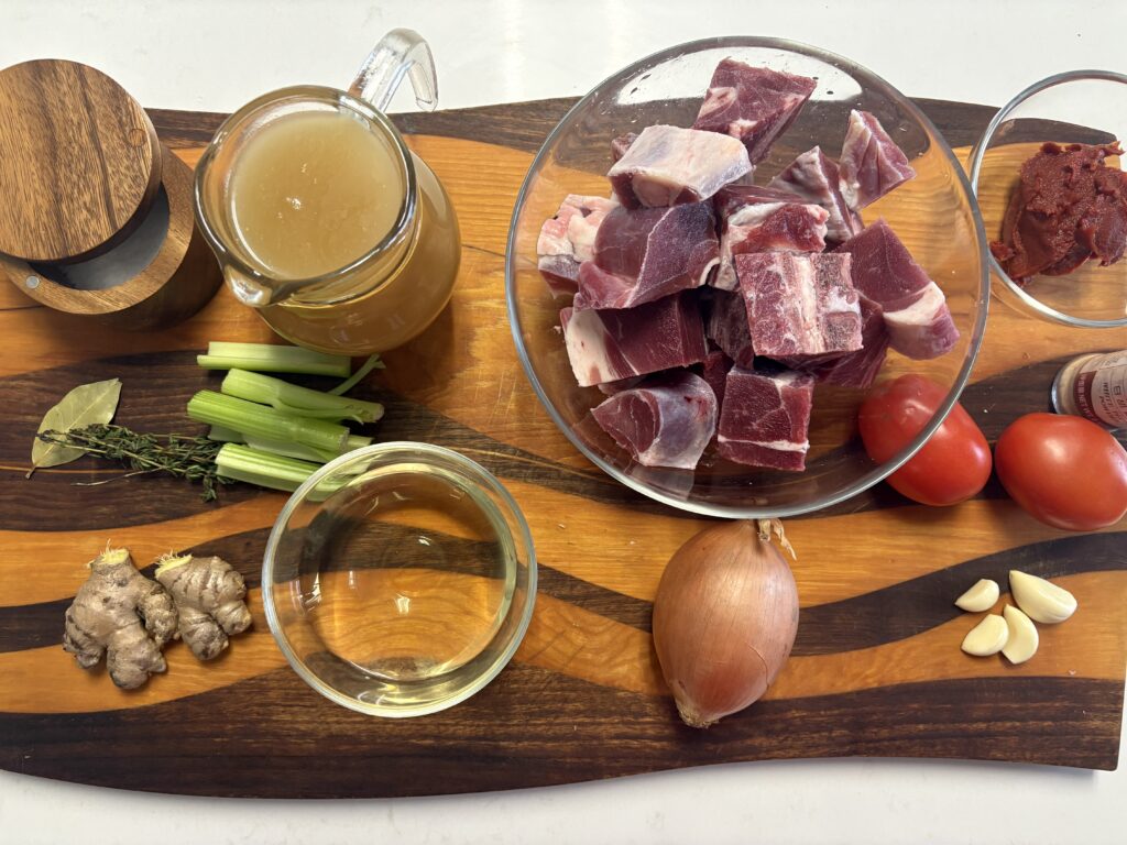 raw goat meat, tomato paste, chicken stock, onions, tomatoes, ginger, garlic, fresh tomatoes, Celery, salt on a wooden board.
