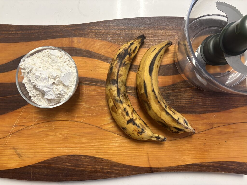 two ripe plantains with flour on the side in a small glass bowl next to a food processor.